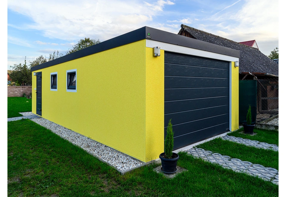 Montovaná garáž 4 x 7 m se žlutou omítkou, garážovými vraty, zateplenými dveřmi, dvěma okny a atikou v antracitové barvě.