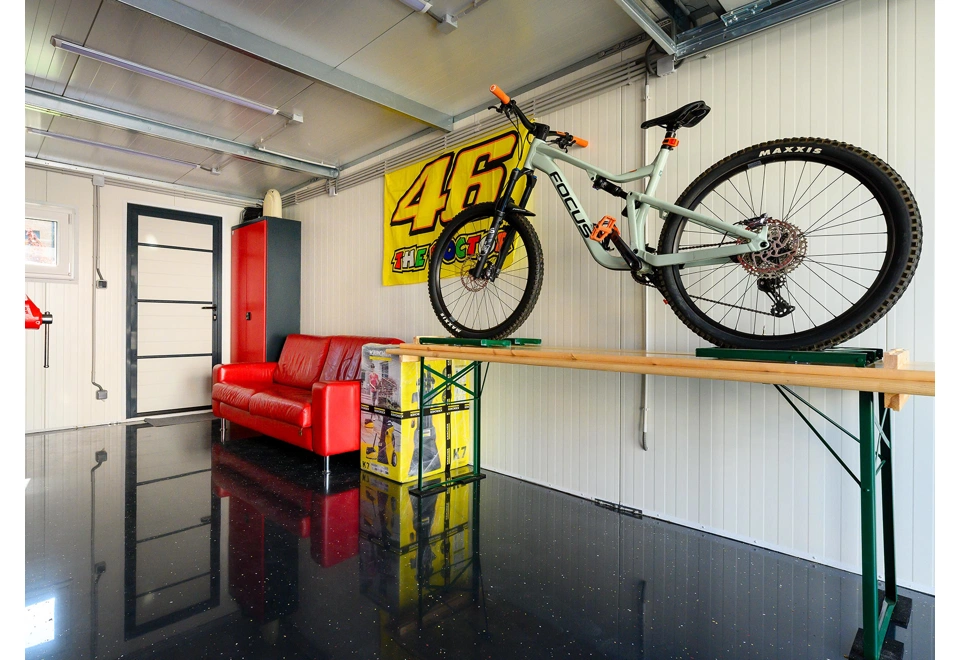 Interiér montované garáže 3 x 7 m s bílými stěnami, oknem a zateplenými dveřmi, gaučem a dílenským stolem.