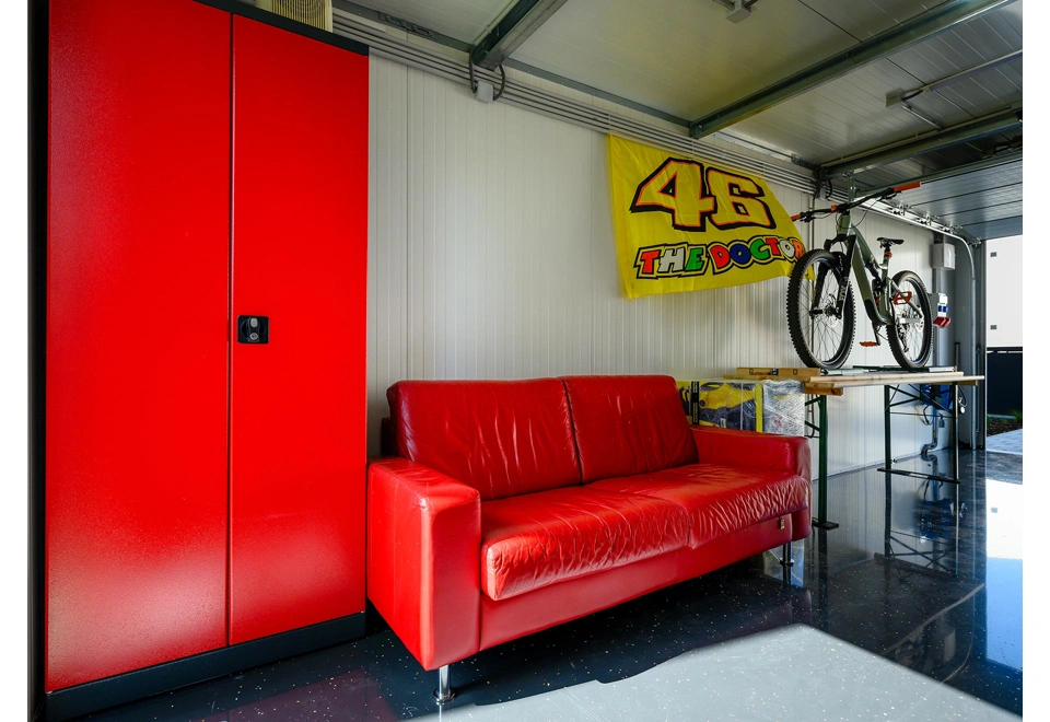 Skříň, gauč a dílenský stůl montované garáže 3 x 7 m s bílými stěnami, garážová sekční vrata u stropu.