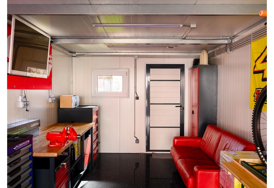 Interiér montované garáže 3 x 7 m s bílými stěnami, oknem a zateplenými dveřmi, gaučem, dílenským stolem a skříňky.