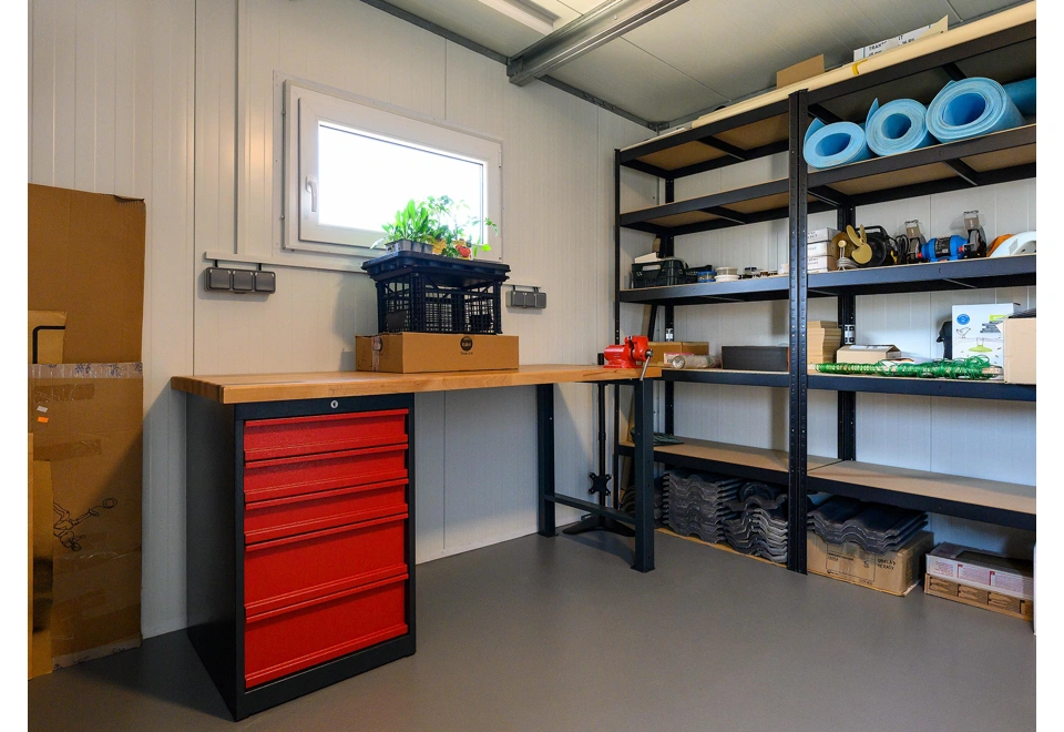 Interiér zahradního domku 4 x 3 m s oknem, dvěma regály a dílenským stolem.